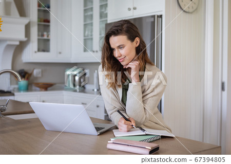 Woman taking notes while using laptop in the kitchen Woman taking notes while using laptop in the kitchen 67394805