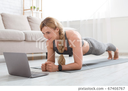 Fitness exercise. Woman in sportswear makes plank on mat and looks at laptop Fitness exercise. Woman in sportswear makes plank on mat and looks at laptop 67408767