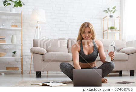 Coach works from home. Smiling woman holds bottle and looks at laptop Coach works from home. Smiling woman holds bottle and looks at laptop 67408796
