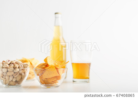 Pistachio, chips and nachos in plates. Glass of unfinished light beer and full bottle of lager Pistachio, chips and nachos in plates. Glass of unfinished light beer and full bottle of lager 67408865