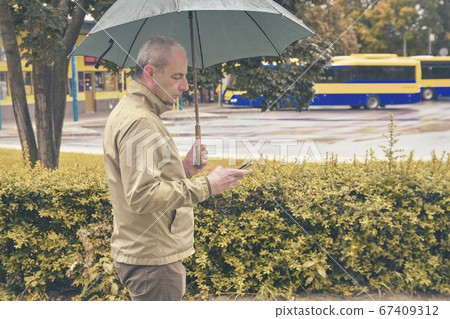 A man standing at the bus station with an umbrella waiting for a bus. Man waiting on a bus, texting on mobile phone in a rainy day. Rainy day in a local town A man standing at the bus station with an umbrella waiting for a bus. Man waiting on a bus, texting on mobile phone in a rainy day. Rainy day in a local town 67409312