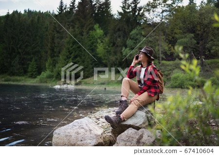 Young woman sitting by lake on a walk outdoors in summer nature, using telephone. Young woman sitting by lake on a walk outdoors in summer nature, using telephone. 67410604