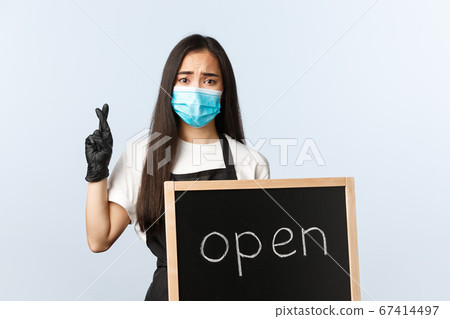 Small business, covid-19 pandemic, preventing virus and employees concept. Hopeful and worried asian female cafe owner, barista hope all okay, cross fingers, showing open sign Small business, covid-19 pandemic, preventing virus and employees concept. Hopeful and worried asian female cafe owner, barista hope all okay, cross fingers, showing open sign 67414497