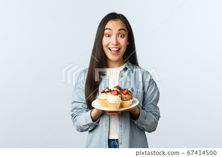 Social distancing lifestyle, covid-19 pandemic, celebrating holidays during coronavirus concept. Excited asian girl holding plate with tasty cupcakes, smiling broadly, invite visit cafe with muffins Social distancing lifestyle, covid-19 pandemic, celebrating holidays during coronavirus concept. Excited asian girl holding plate with tasty cupcakes, smiling broadly, invite visit cafe with muffins 67414500