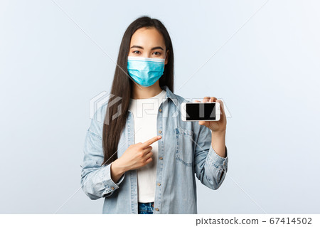 Social distancing lifestyle, covid-19 pandemic and technology concept. Cute asian woman in medical mask pointing finger at smartphone display, promote application or game Social distancing lifestyle, covid-19 pandemic and technology concept. Cute asian woman in medical mask pointing finger at smartphone display, promote application or game 67414502
