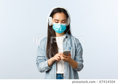 Social distancing lifestyle, covid-19 pandemic and self-isolation leisure concept. Modern cute asian girl in medical mask, listening music wireless headphones, using mobile phone app Social distancing lifestyle, covid-19 pandemic and self-isolation leisure concept. Modern cute asian girl in medical mask, listening music wireless headphones, using mobile phone app 67414504