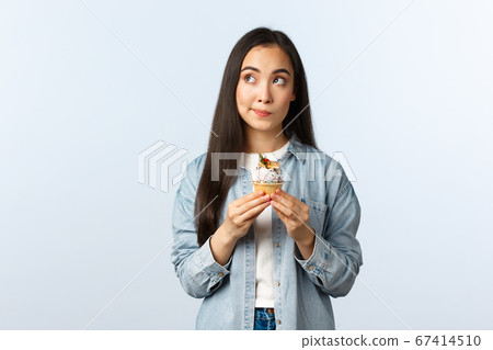 Social distancing lifestyle, covid-19 pandemic, celebrating holidays during coronavirus concept. Thoughtful cute asian girl deciding eat cupcake or note, look up thinking, holding delicious dessert Social distancing lifestyle, covid-19 pandemic, celebrating holidays during coronavirus concept. Thoughtful cute asian girl deciding eat cupcake or note, look up thinking, holding delicious dessert 67414510