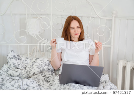 woman drinks tea and eats in bed at computer home. 67431557