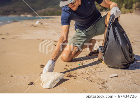 Man in gloves pick up plastic bags that pollute sea. Problem of spilled rubbish trash garbage on the beach sand caused by man-made pollution and environmental, campaign to clean volunteer in concept Man in gloves pick up plastic bags that pollute sea. Problem of spilled rubbish trash garbage on the beach sand caused by man-made pollution and environmental, campaign to clean volunteer in concept 67434720