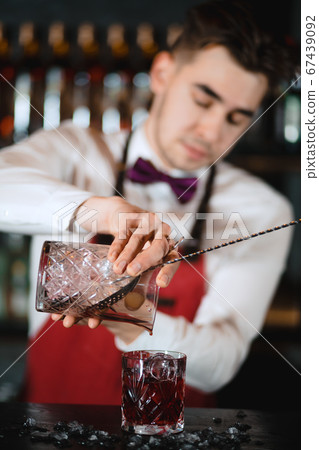 Barman holding a long spoon and glass filled with ice cubes on the bar counter 67439092