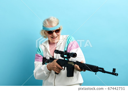 Senior woman in sports wear and black sanglasses with a rifle isolated on blue 67442731