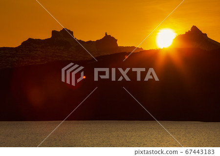 Beautiful sunset in rocky mountains and lake shore, red orange sun sets behind silhouette of cliff mountain range. Natural lens flare in sky, sunlight leaks. 67443183