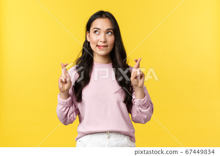 People emotions, lifestyle and fashion concept. Dreamy and hopeful cute asian girl having aspirations, biting lip and looking upper left corner tempting, cross fingers good luck, yellow background 67449834