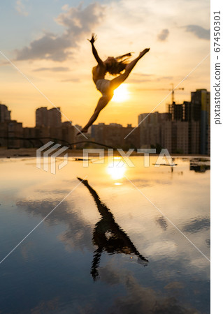 Female flexible dancer jumping during sunset on cityscape background with reflection in the water. Concept of freedom and happiness  67450301