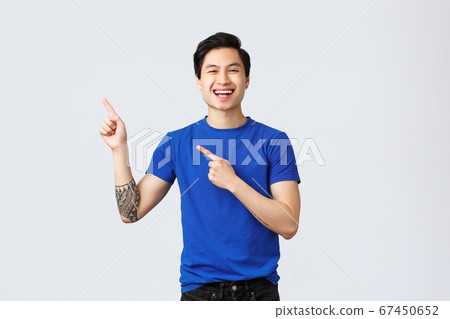 Different emotions, people lifestyle and advertising concept. Handsome carefree asian man in blue t-shirt, point fingers upper left corner and smiling delighted, introduce good new promo, show banner Different emotions, people lifestyle and advertising concept. Handsome carefree asian man in blue t-shirt, point fingers upper left corner and smiling delighted, introduce good new promo, show banner 67450652
