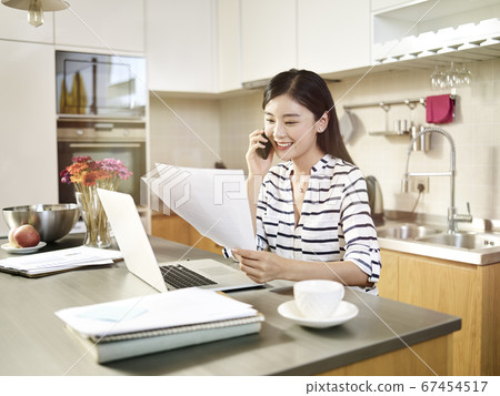 young asian woman sitting at kitchen counter working using laptop computer talking on cellphone. young asian woman sitting at kitchen counter working using laptop computer talking on cellphone. 67454517