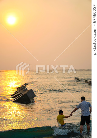 View from the beach with a yellow glowing sun setting in the horizon with an Asian family playing in the water. 67460057