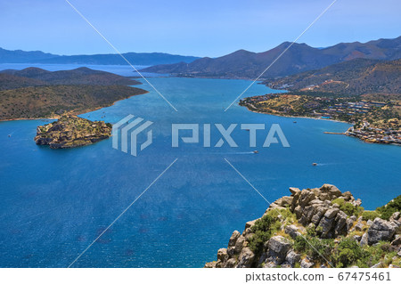 View of harbor between Spinalonga island and peninsula, known as Kalydon, and Plaka village from above and rocky slope in foreground, Crete, Greece. View of harbor between Spinalonga island and peninsula, known as Kalydon, and Plaka village from above and rocky slope in foreground, Crete, Greece. 67475461
