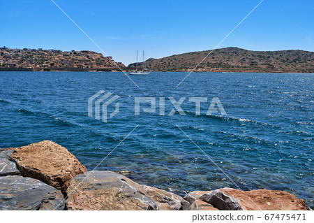 View of Spinalonga island lagoon and Venetian fortress with anchored yacht and breakwater stones in foreground, Crete, Greece View of Spinalonga island lagoon and Venetian fortress with anchored yacht and breakwater stones in foreground, Crete, Greece 67475471