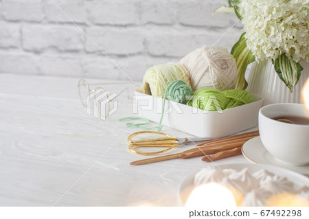 Green yarn in a white plastic basket. Knitting needles and scissors. Tea in a cup and biz. Aged light brick background. Needlework and female hobby concept. 67492298