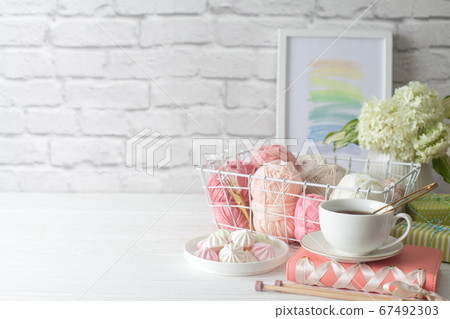 Needlework accessories in a white metal basket. Pink yarn. Gray and wooden knitting needles. White flowers in a vase. Background old brick. 67492303