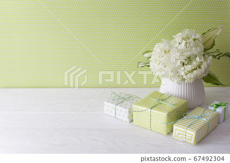 Little green gifts and a bouquet of white hydrangea in a vase. Green wall and white wooden table. Little green gifts and a bouquet of white hydrangea in a vase. Green wall and white wooden table. 67492304