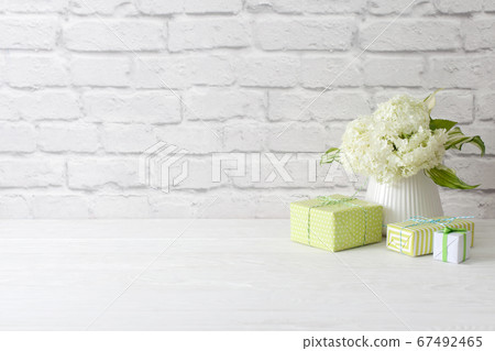 Little green gifts and a bouquet of white hydrangea in a vase. brick wall and white wooden table. 67492465