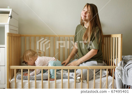 Young tired mother in casual clothes sitting together with her baby girl in a cot, real motherhood 67495401