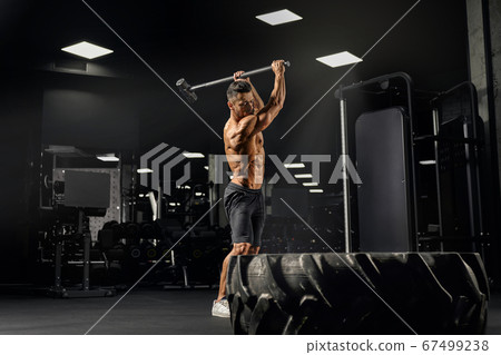 Muscular man hitting giant wheel with sledgehammer. Muscular man hitting giant wheel with sledgehammer. 67499238