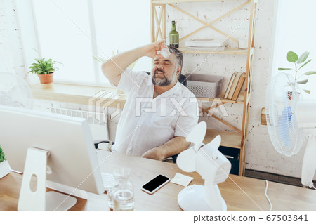 Businessman, manager in office with computer and fan cooling off, feeling hot, flushed 67503841