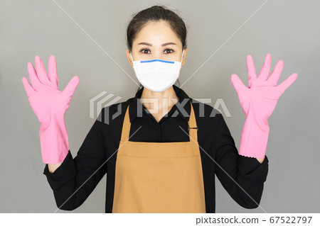Young woman housewife in brown apron and pink gloves wore medical face mask,spread her fingers and raise both hands isolated on gray background,housework and household concept. 67522797