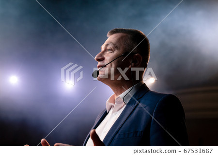 Showman. Middle Age Male entertainer, presenter or actor on stage. Arms to sides, smoke on background of spotlight. Front view portrait of a male public speaker speaking at the microphone, pointing Showman. Middle Age Male entertainer, presenter or actor on stage. Arms to sides, smoke on background of spotlight. Front view portrait of a male public speaker speaking at the microphone, pointing 67531687