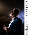 Showman. Middle Age Male entertainer, presenter or actor on stage. Arms to sides, smoke on background of spotlight. Front view portrait of a male public speaker speaking at the microphone, pointing 67531693