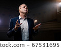 Showman. Middle Age Male entertainer, presenter or actor on stage. Arms to sides, smoke on background of spotlight. Front view portrait of a male public speaker speaking at the microphone, pointing 67531695