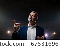 Showman. Middle Age Male entertainer, presenter or actor on stage. Arms to sides, smoke on background of spotlight. Front view portrait of a male public speaker speaking at the microphone, pointing 67531696