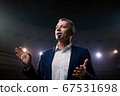 Showman. Middle Age Male entertainer, presenter or actor on stage. Arms to sides, smoke on background of spotlight. Front view portrait of a male public speaker speaking at the microphone, pointing 67531698