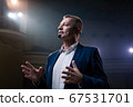 Showman. Middle Age Male entertainer, presenter or actor on stage. Arms to sides, smoke on background of spotlight. Front view portrait of a male public speaker speaking at the microphone, pointing 67531701