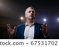Showman. Middle Age Male entertainer, presenter or actor on stage. Arms to sides, smoke on background of spotlight. Front view portrait of a male public speaker speaking at the microphone, pointing 67531702