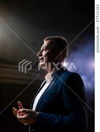 Showman. Middle Age Male entertainer, presenter or actor on stage. Arms to sides, smoke on background of spotlight. Front view portrait of a male public speaker speaking at the microphone, pointing 67532163