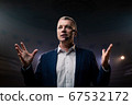 Showman. Middle Age Male entertainer, presenter or actor on stage. Arms to sides, smoke on background of spotlight. Front view portrait of a male public speaker speaking at the microphone, pointing 67532172
