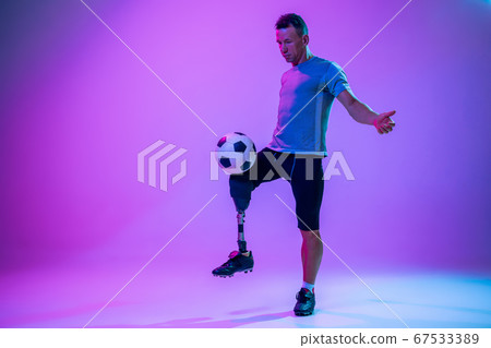 Athlete with disabilities or amputee isolated on gradient studio background. Professional male football player with leg prosthesis training and practicing in studio. Athlete with disabilities or amputee isolated on gradient studio background. Professional male football player with leg prosthesis training and practicing in studio. 67533389