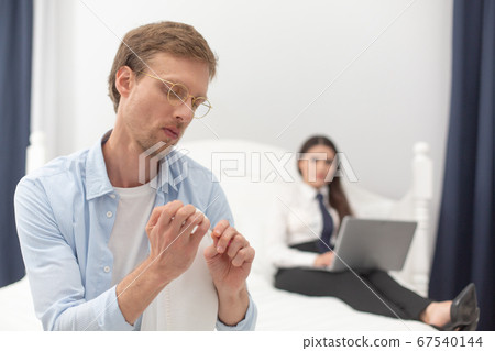 Househusband polishing his nails. Man acting feminine by polishing his nails while his wife is working with computer in background. Selective focus on man in foreground Househusband polishing his nails. Man acting feminine by polishing his nails while his wife is working with computer in background. Selective focus on man in foreground 67540144