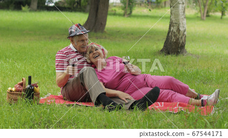 Family weekend picnic in park. Active senior old caucasian couple sit on blanket and drink wine Family weekend picnic in park. Active senior old caucasian couple sit on blanket and drink wine 67544171