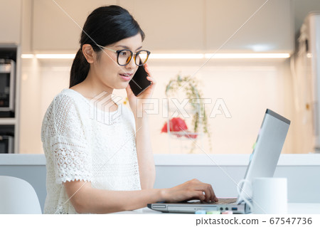 在家工作一名戴著眼鏡的女人在餐廳的筆記本電腦前戴著智能手機 在家工作一名戴著眼鏡的女人在餐廳的筆記本電腦前戴著智能手機 67547736