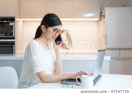 在家工作一名戴著眼鏡的女人在餐廳的筆記本電腦前戴著智能手機 在家工作一名戴著眼鏡的女人在餐廳的筆記本電腦前戴著智能手機 67547739