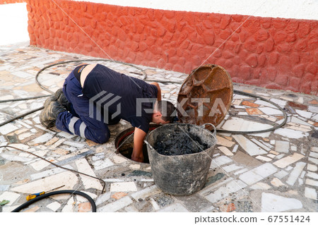 Worker cleans the drains hatch and removes dirt and debris from the sewer. Plumber cleans the sewer 67551424
