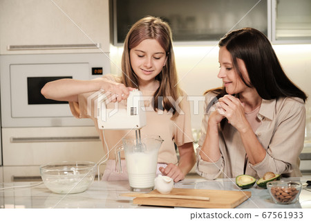 Happy blond teenage girl holding electric mixer in large jug with fresh milk Happy blond teenage girl holding electric mixer in large jug with fresh milk 67561933