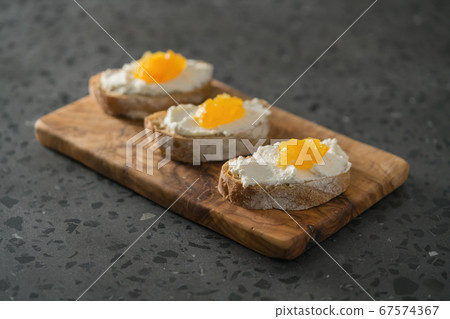 Three ciabatta slices with ricotta and orange marmalade on olive wood board on terrazzo surface 67574367