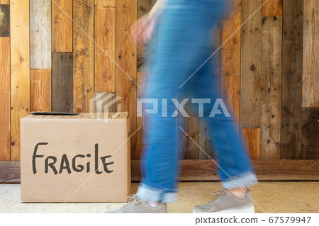 Young woman walks with moving box in new home near wooden wall, holding cardboard moving boxes on moving day retro design fragile written on box 67579947