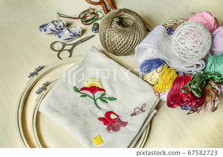 Light wooden table with embroidery in a round frame, colored threads and scissors Light wooden table with embroidery in a round frame, colored threads and scissors 67582723